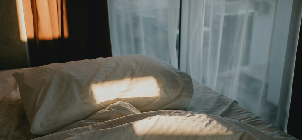 Lit défait avec la lumière du soleil sur l’oreiller et la couverture devant une fenêtre aux rideaux blancs, le matin.