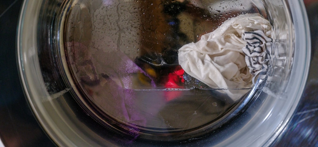 Nahaufnahme einer Waschmaschinentrommel, in der sich nasse, weiße und farbige Kleidung befindet. Es sind Wassertropfen auf der Innenseite der Trommel sichtbar.