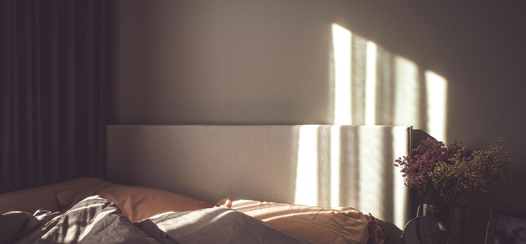 The picture shows a bed with two pillows and blankets, illuminated by a ray of sunlight. Next to the bed is a bedside table with a mirror, a picture and flowers. The room looks cosy and inviting.