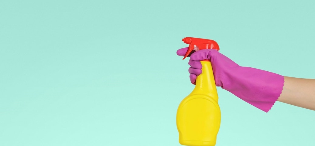 Eine Hand mit einem pinken Handschuh hält eine gelbe Sprühflasche mit rotem Deckel vor einem türkisfarbenen Hintergrund.
