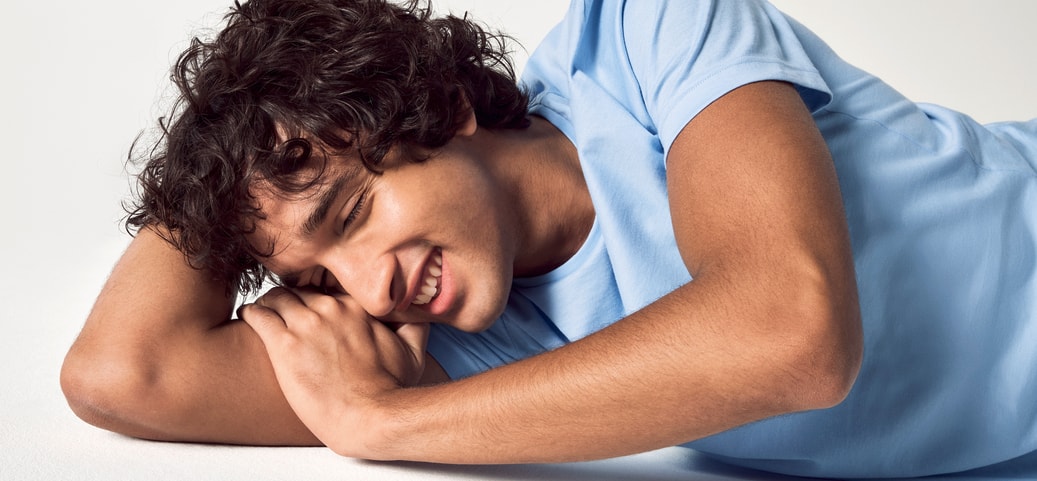 The picture shows a smiling man with curly hair lying on his side on a white background and wearing a light blue T-shirt from CALIDA. He has his eyes closed, symbolising laughter in his sleep.
