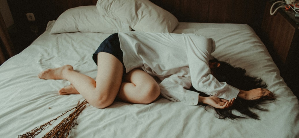 The picture shows a woman lying on a bed without a pillow in a dimly lit room.