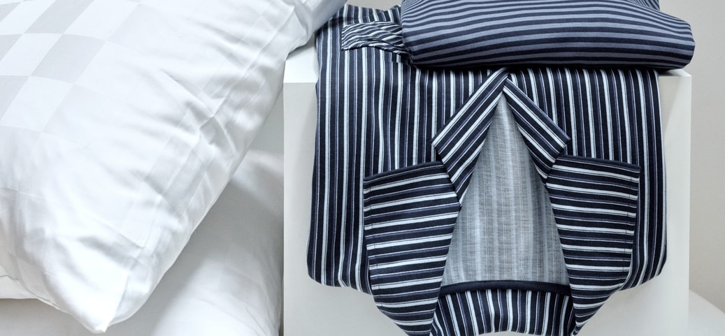 Zusammengefalteter gestreifter Schlafanzug in Blau- und Grautönen auf einem weißen Tisch neben einem weißen Kissen mit dezentem Karomuster. Im Hintergrund hängt ein schwarzes Kleidungsstück.