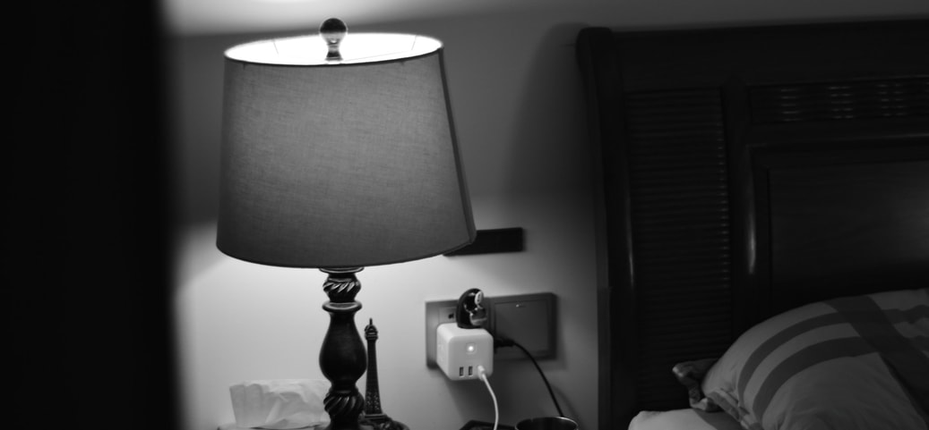 Nachttisch mit Lampe, Ladegerät in der Steckdose, Metallbecher und Smartphone neben einem Bett mit Kopfkissen.
