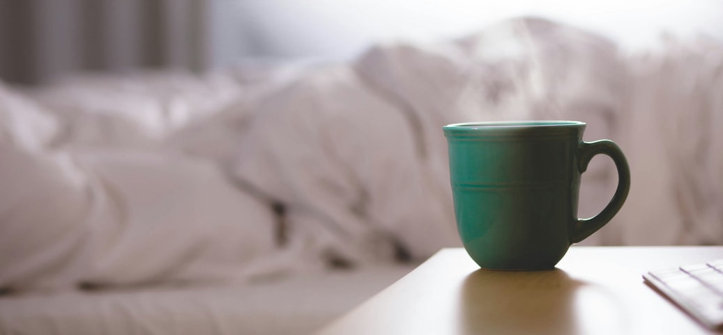 A green cup of steaming coffee stands on a bedside table in front of an unmade bed, symbolising coffee before sleep.