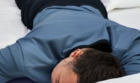 Person lying on their stomach on a white bed, sleeping, wearing a blue shirt and dark trousers – calm sleep situation