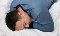 Man sleeping on his side on a white bed, wearing blue CALIDA sleepwear; symbolic image for restorative REM sleep.