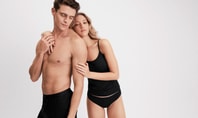 Woman wearing a black spaghetti‑strap top and matching brief from CALIDA’s SleekSkin series standing next to a man wearing a black boxer brief from CALIDA’s Natural Cooling series against a light background