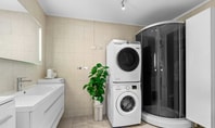 Bagno moderno con lavandino, specchio illuminato, lavatrice e asciugatrice impilate, doccia in vetro e grande vaso di piante su piastrelle chiare