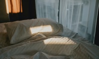 Letto sfatto con luce del sole su cuscino e coperta davanti a una finestra con tende bianche al mattino.