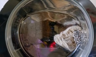 Nahaufnahme einer Waschmaschinentrommel, in der sich nasse, weiße und farbige Kleidung befindet. Es sind Wassertropfen auf der Innenseite der Trommel sichtbar.