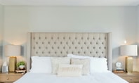 Bright bedroom with beige, upholstered headboard, two bedside tables, golden lamps and white bed linen - symbolising dry air in the bedroom.