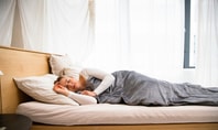  Auf dem Bild liegt eine Frau seitlich im Bett, ihre Augen sind geschlossen und ihr Gesicht zeigt Anzeichen von Entspannung. Die Frau ist von einer grauen Decke bedeckt, die sanft über ihren Körper drapiert ist. Unter ihrem Kopf liegt ein weißes Kissen, das eine weiche Unterstützung bietet. Die Szene strahlt Ruhe und Gelassenheit aus, während die Frau in einem Moment der Ruhe und des Friedens verweilt.