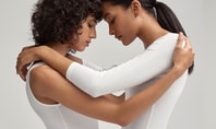 Two women in plain white bodysuits. The left half of the picture shows the two women in an intimate embrace, while the right half shows one of the women posing confidently with one hand behind her head.