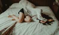 Auf dem Bild ist eine Frau zu sehen, die auf einem Bett ohne Kissen in einem dämmrigen Raum liegt.
