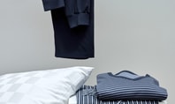 Zusammengefalteter gestreifter Schlafanzug in Blau- und Grautönen auf einem weißen Tisch neben einem weißen Kissen mit dezentem Karomuster. Im Hintergrund hängt ein schwarzes Kleidungsstück.