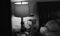 Nachttisch mit Lampe, Ladegerät in der Steckdose, Metallbecher und Smartphone neben einem Bett mit Kopfkissen.