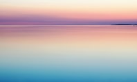 Ein beruhigender Sonnenaufgang über einem stillen Ozean, mit sanften Farbübergängen von Blau über Rosa zu hellem Gelb, die den Himmel und das Wasser harmonisch verschmelzen lassen.