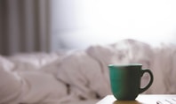A green cup of steaming coffee stands on a bedside table in front of an unmade bed, symbolising coffee before sleep.