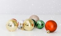 Baubles natalizi in vari colori, tra cui oro, rosso, verde, argento e glitter su sfondo bianco, con un sottile effetto fiocco di neve.