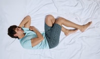 Uomo sdraiato su un letto bianco che dorme di lato, indossa una maglietta azzurra e pantaloncini a fantasia - scena della campagna CALIDA sleep through.
