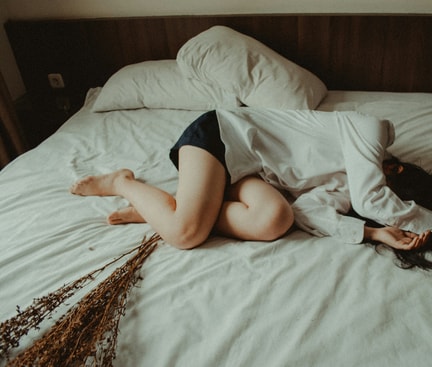 Person liegt zusammengerollt auf einem Bett, trägt Hemd und Shorts.