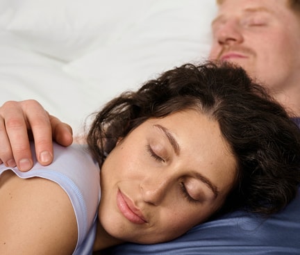 Two people sleeping closely snuggled together on a white bed – a typical couple sleeping position.