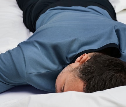 Person lying on their stomach on a white bed, sleeping, wearing a blue shirt and dark trousers – calm sleep situation