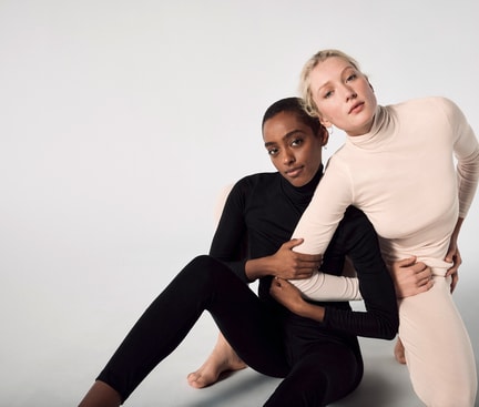 Two people leaning closely together in black and beige turtleneck outfits against a light background.