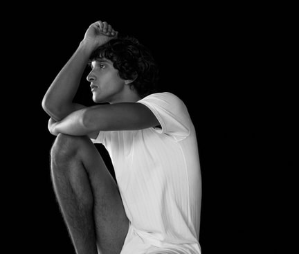 Person in a white T‑shirt sitting thoughtfully against a black background – minimalist, contemplative portrait scene