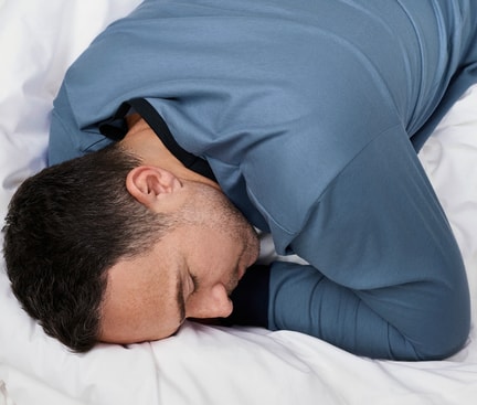 Man sleeping on his side on a white bed, wearing blue CALIDA sleepwear; symbolic image for restorative REM sleep.