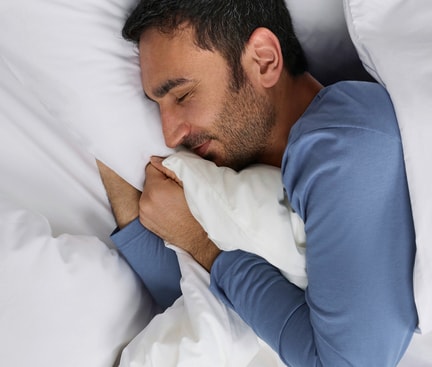Schlafender Mann in blauer Nachtwäsche in weissem Bett; Symbolbild für erholsamen Schlaf und ein starkes Immunsystem