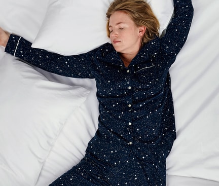 Personne endormie en pyjama bleu foncé à motifs d'étoiles sur un lit blanc ; image symbolique pour le sujet de sensation de froid et de sueur en même temps pendant la nuit