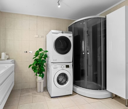 Modern bathroom with sink, illuminated mirror, stacked washing machine and dryer, glass shower and large plant pot on light tiles