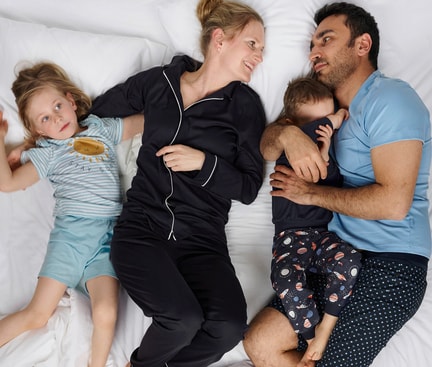 Family lying together on a white bed; two adults and two children wearing CALIDA sleepwear, relaxed bedtime scene illustrating why sleeping in pyjamas is beneficial.