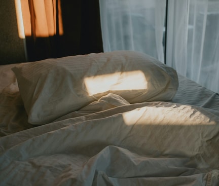 Unordentliches Bett mit Sonnenlicht auf Kissen und Decke vor einem Fenster mit weißen Vorhängen am Morgen.