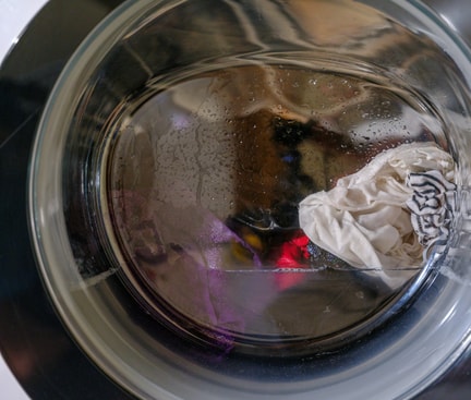 Nahaufnahme einer Waschmaschinentrommel, in der sich nasse, weiße und farbige Kleidung befindet. Es sind Wassertropfen auf der Innenseite der Trommel sichtbar.