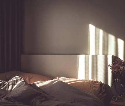Das Bild zeigt ein Bett mit zwei Kissen und Decken, von einem Sonnenstrahl beleuchtet. Neben dem Bett steht ein Nachttisch mit einem Spiegel, einem Bild und Blumen. Der Raum wirkt gemütlich und einladend.