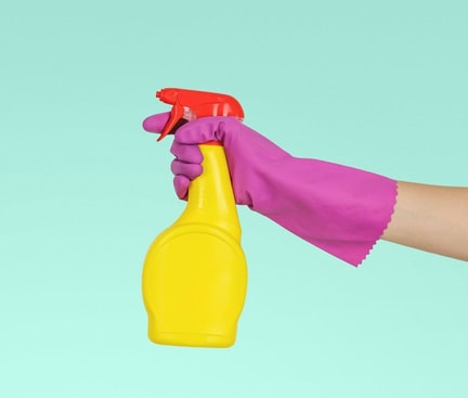 Eine Hand mit einem pinken Handschuh hält eine gelbe Sprühflasche mit rotem Deckel vor einem türkisfarbenen Hintergrund.