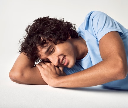 Das Bild zeigt einen lächelnden Mann mit lockigem Haar, der seitlich auf einem weißen Untergrund liegt und ein hellblaues T-Shirt von CALIDA trägt. Er hat die Augen geschlossen, was das Lachen im Schlaf symbolisieren soll.