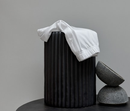 Das Bild zeigt ein weißes Kleidungsstück, das über einem schwarzen, zylinderförmigen Objekt drapiert ist. Daneben befinden sich zwei graue, schalenförmige Objekte, die übereinander gestapelt sind.