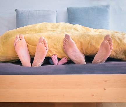 Eltern und Baby beim Co-Sleeping, ihre Füße schauen gemeinsam unter der Bettdecke hervor.