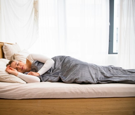  Auf dem Bild liegt eine Frau seitlich im Bett, ihre Augen sind geschlossen und ihr Gesicht zeigt Anzeichen von Entspannung. Die Frau ist von einer grauen Decke bedeckt, die sanft über ihren Körper drapiert ist. Unter ihrem Kopf liegt ein weißes Kissen, das eine weiche Unterstützung bietet. Die Szene strahlt Ruhe und Gelassenheit aus, während die Frau in einem Moment der Ruhe und des Friedens verweilt.