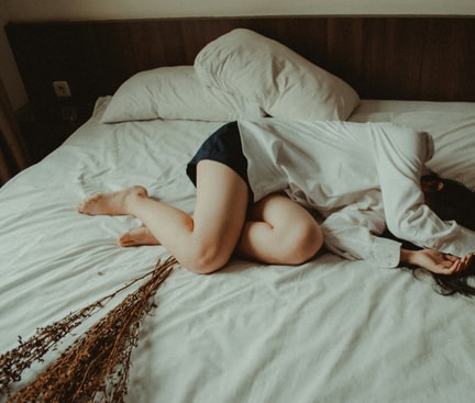Auf dem Bild ist eine Frau zu sehen, die auf einem Bett ohne Kissen in einem dämmrigen Raum liegt.