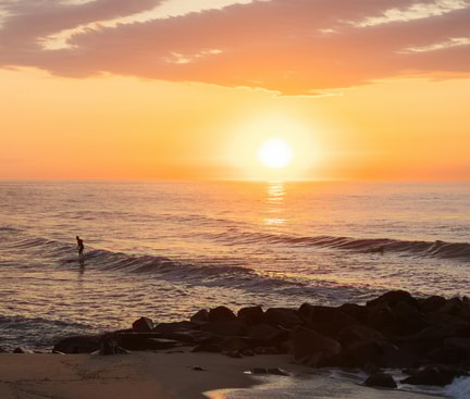 Früher Morgen am Strand, eine Person steht im Meer, während die Sonne gerade aufgeht.