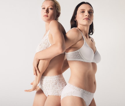Auf dem Bild stehen zwei Frauen Rücken an Rücken und seitlich zur Kamera. Sie tragen weiße Unterwäsche von CALIDA. Die Kleidung betont ihre Körperkonturen, während sie eine elegante Haltung einnehmen. Der Hintergrund ist in einem sanften Weißgrau gehalten.