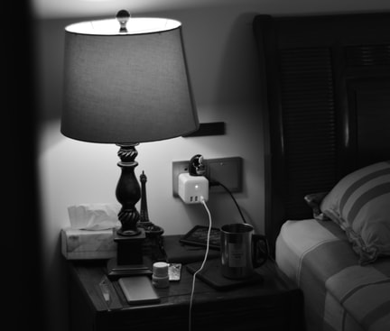 Nachttisch mit Lampe, Ladegerät in der Steckdose, Metallbecher und Smartphone neben einem Bett mit Kopfkissen.