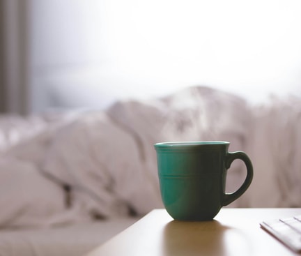 Eine grüne Tasse mit dampfendem Kaffee steht auf einem Nachttisch vor einem ungemachten Bett, Symbol für Kaffee vor dem Schlafen.