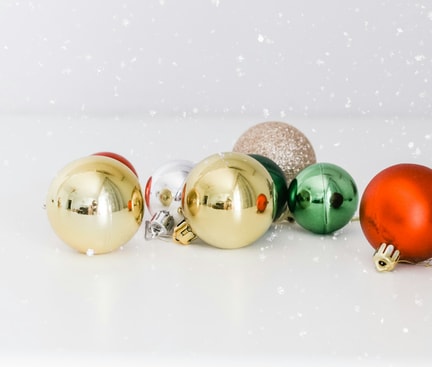 Weihnachtskugeln in verschiedenen Weihnachtsfarben, darunter Gold, Rot, Grün, Silber und Glitzer auf einem weißen Hintergrund, mit einem dezenten Schneeflocken-Effekt.