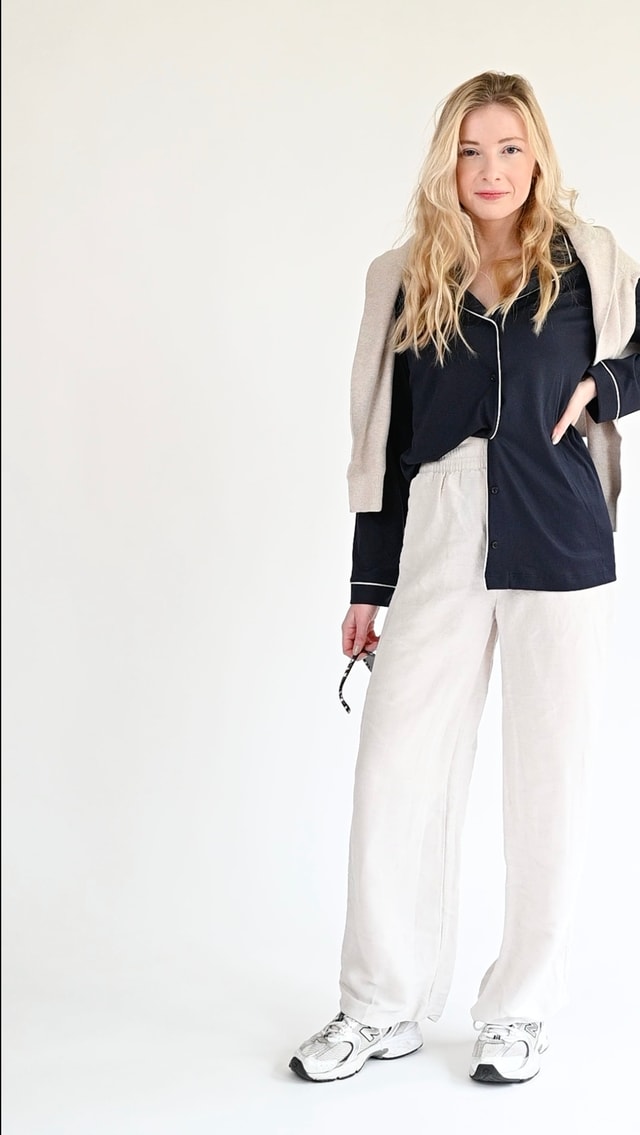 Eine Frau trägt ein lässiges Oberteil im CALIDA-Pyjama-Stil, kombiniert mit einer hellen Hose, einer beigen Strickjacke, die sie sich über die Schultern gelegt hat, und Turnschuhen - der perfekte After-Work-Loungewear-Look.